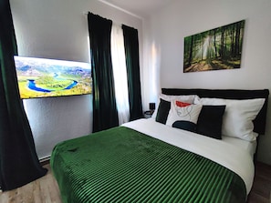 Et hotelværelse med en seng, et skovmotiv på en væg, et landskabsmaleri og et vindue med gardiner.