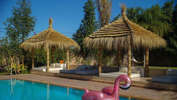 A swimming pool with a pink flamingo float, surrounded by thatched umbrellas and lounge areas. - Villa Atika Maison d'hôtes & SPA (Marrakech)