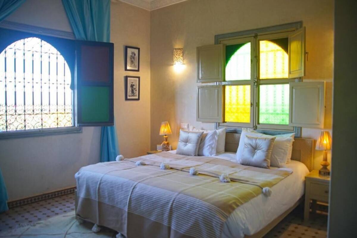 A neatly made bed with white linens and pillows, a bedside lamp, and a window with curtains.