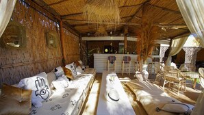 A room with a thatched roof, wooden walls, and a long sofa with patterned cushions. There is a small bar area in the background. - Villa Atika Maison d'hôtes & SPA (Marrakech)