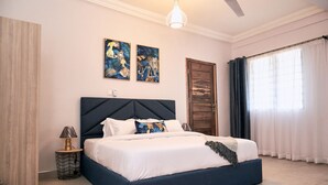 A bedroom with a large bed, a ceiling fan, two wall art pieces, a wooden door, and a window with curtains. - Red Lotus Oasis (Accra)