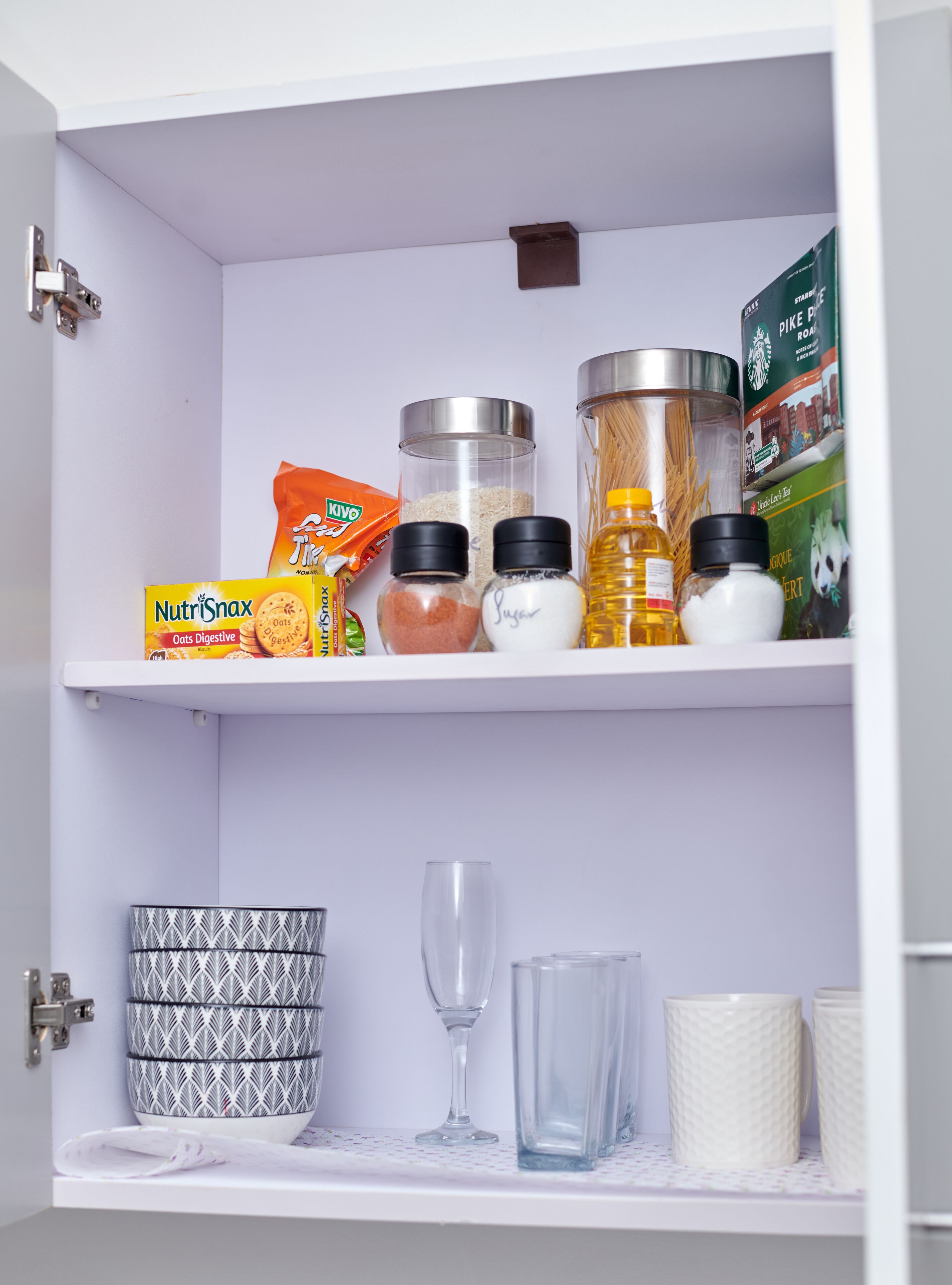 Una despensa con estantes que contienen diversos alimentos, un cuenco estampado, una copa de vino y un juego de tazas blancas.