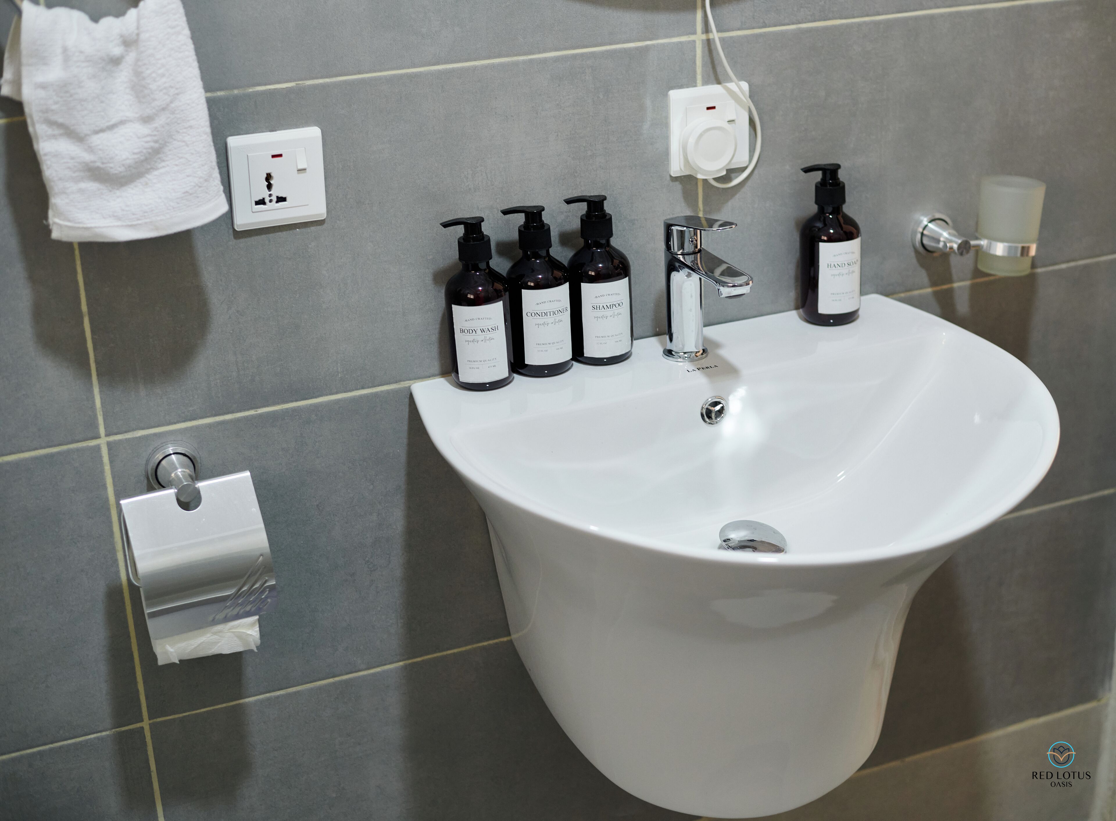 Un baño con un lavabo blanco, dispensadores de jabón negros, una botella de jabón para manos, un dispensador de jabón de vidrio y un dispensador de toallas de papel.