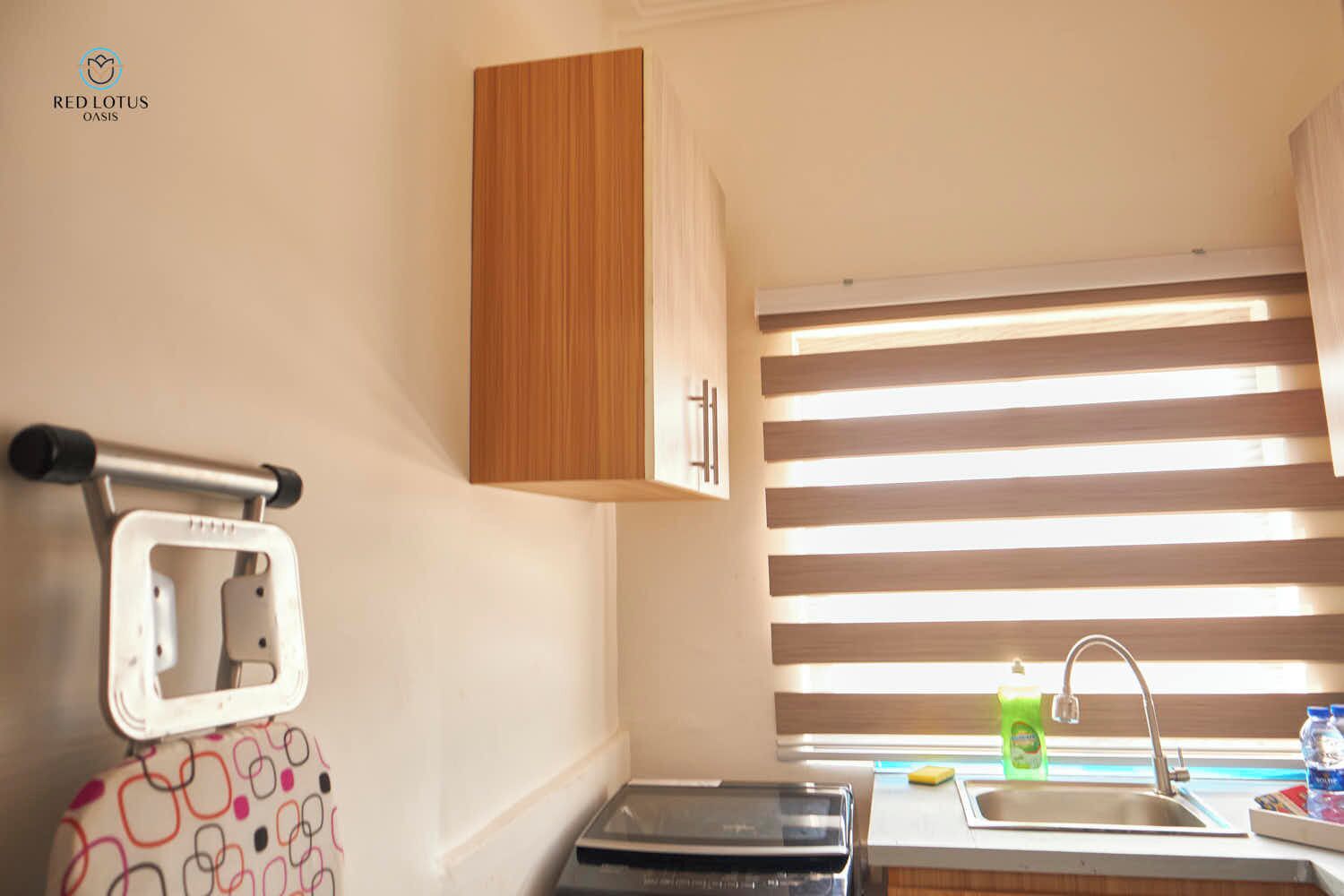 A compact kitchen with a wooden cabinet, a built-in microwave, a sink, and a window with horizontal blinds.