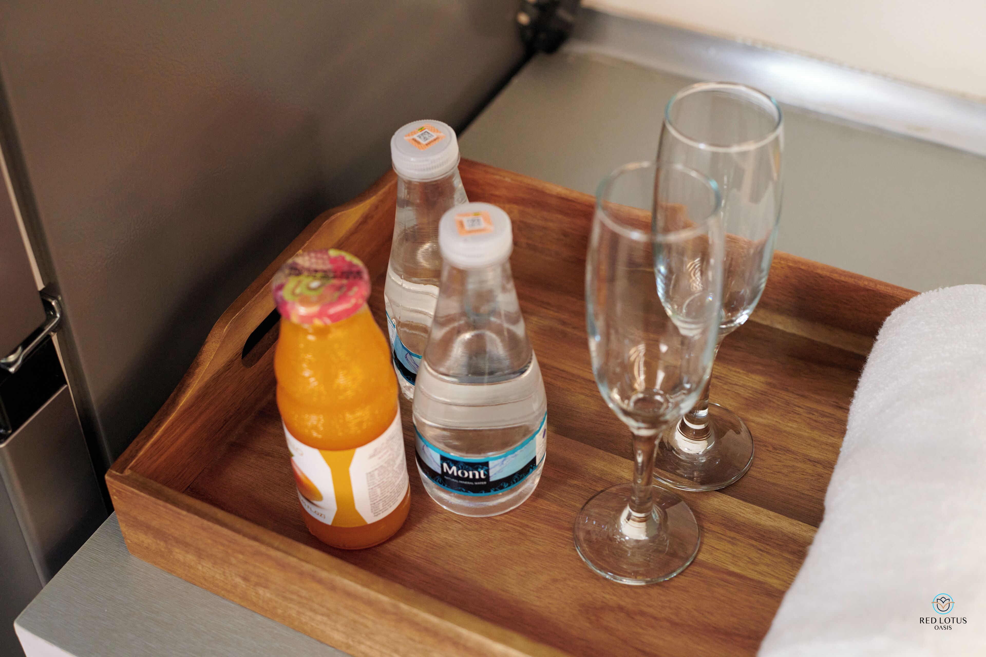 A wooden tray with a bottle of orange juice, two water bottles, and a wine glass on a countertop.