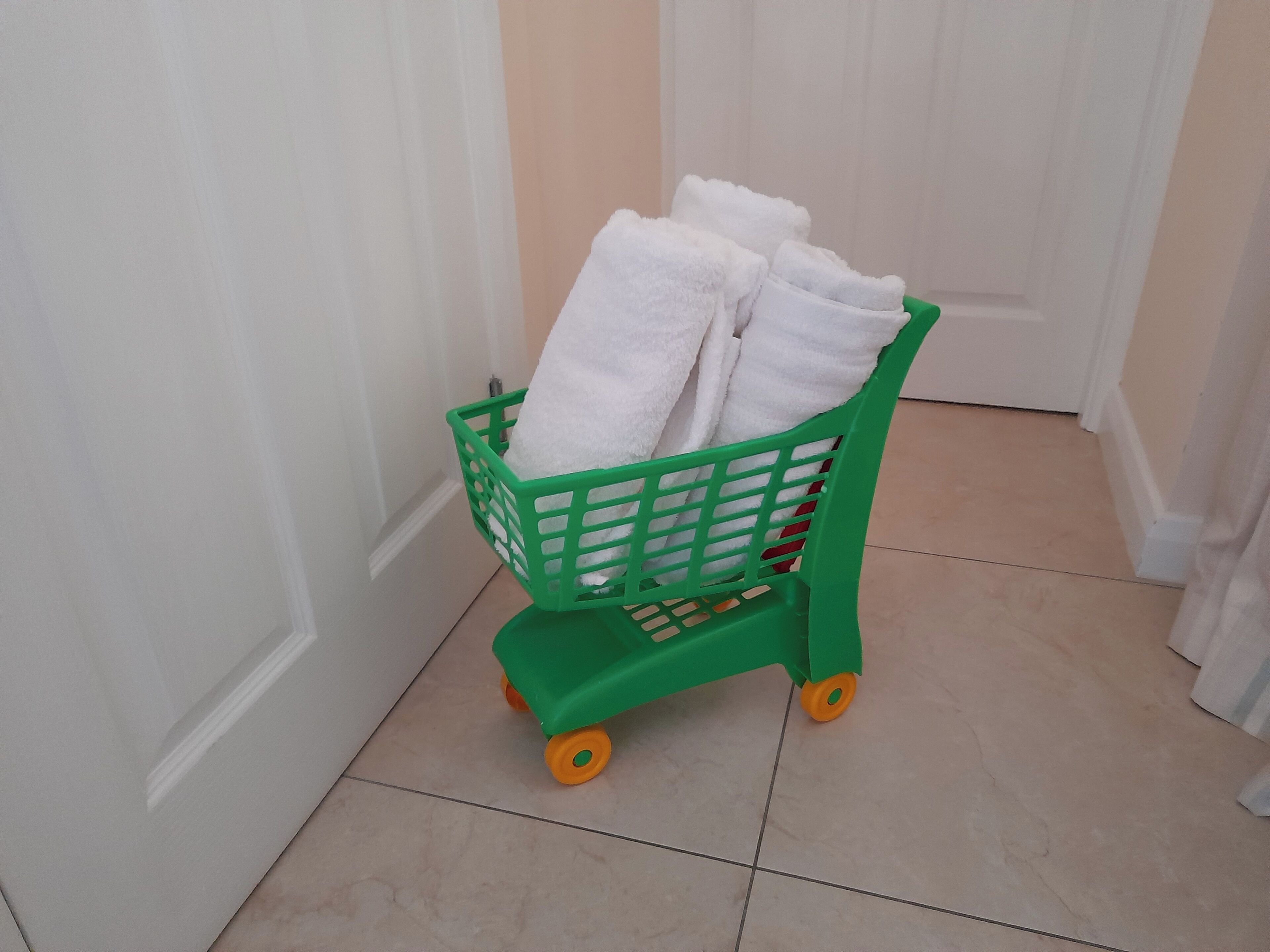 A green toy cart with white towels inside, placed on a tiled floor near a white door.