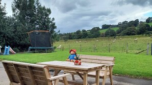 Un banco de madera con una mesa en un parque con un trampolín y un campo cubierto de césped con ovejas al fondo.
