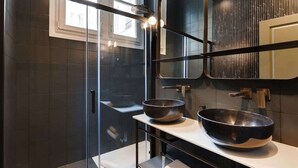 A modern bathroom with dark tiles, a glass shower enclosure, and two large bowls on a white countertop.