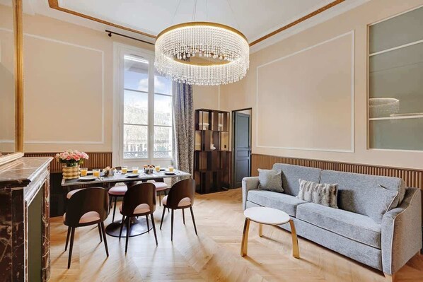 A spacious living room with a grey sofa, a dining table with chairs, a chandelier, and a large window.
