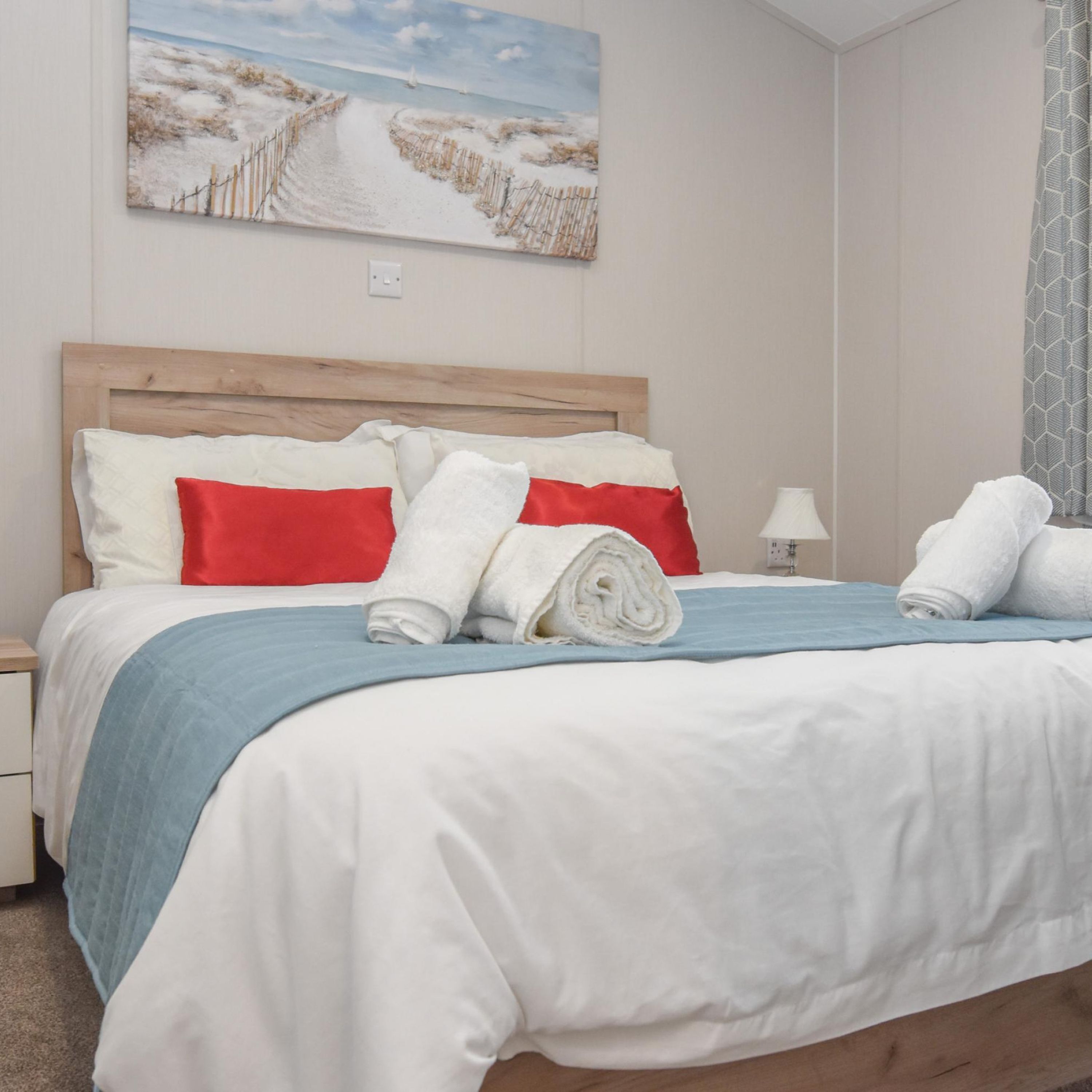 A neatly made bed with white and red pillows, a blue blanket, and rolled towels.