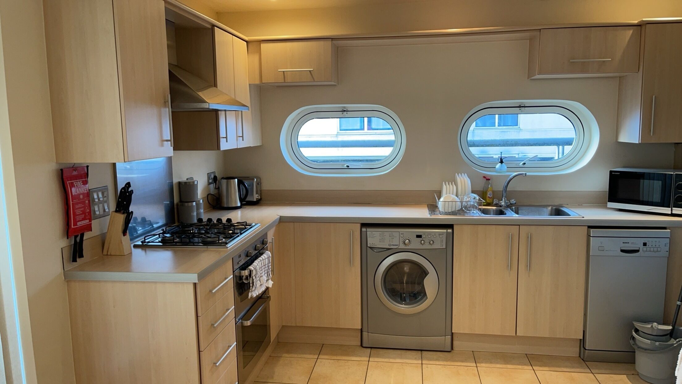 A compact kitchen with a stove, microwave, sink, and washing machine. There are two windows providing natural light.