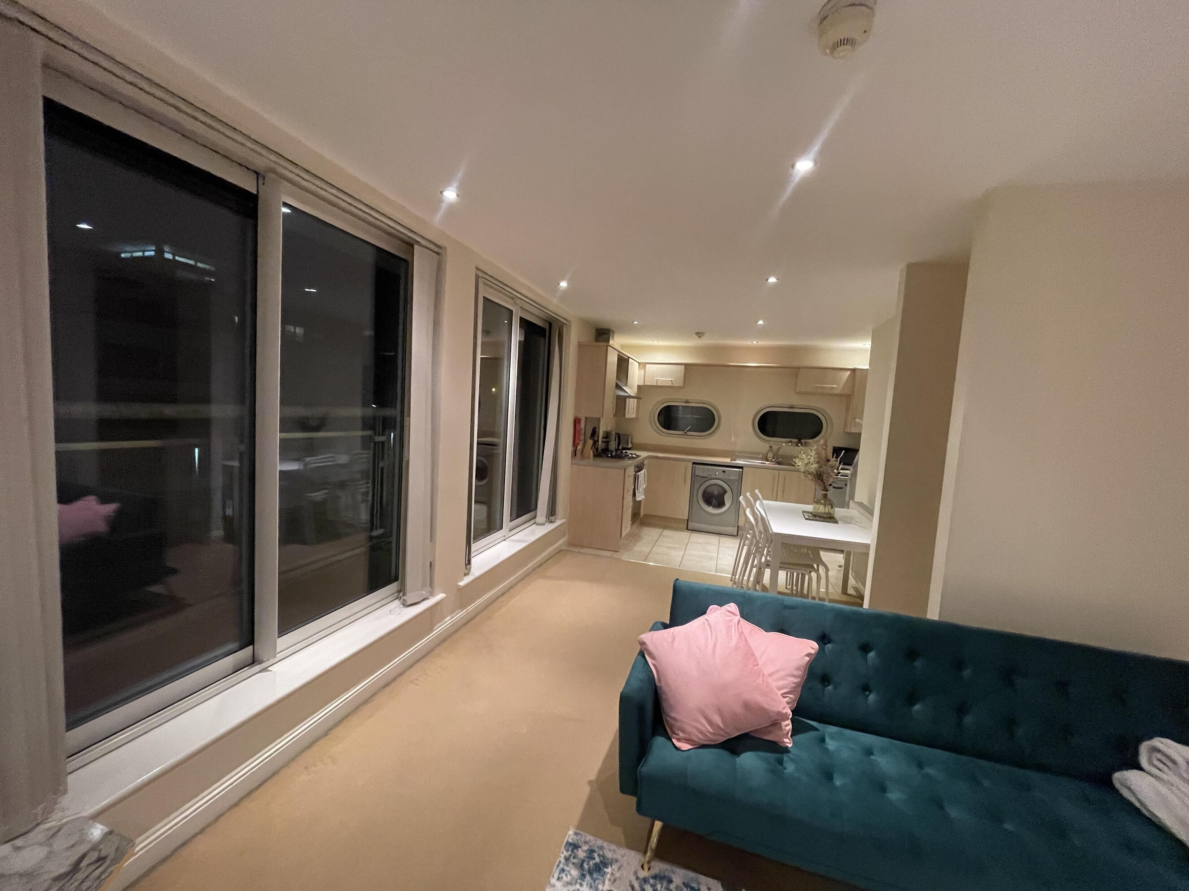 Un apartamento moderno con una sala de estar que presenta un sofá color esmeralda y una almohada rosa, una cocina con lavadora y ventanas amplias que dejan entrar luz natural.