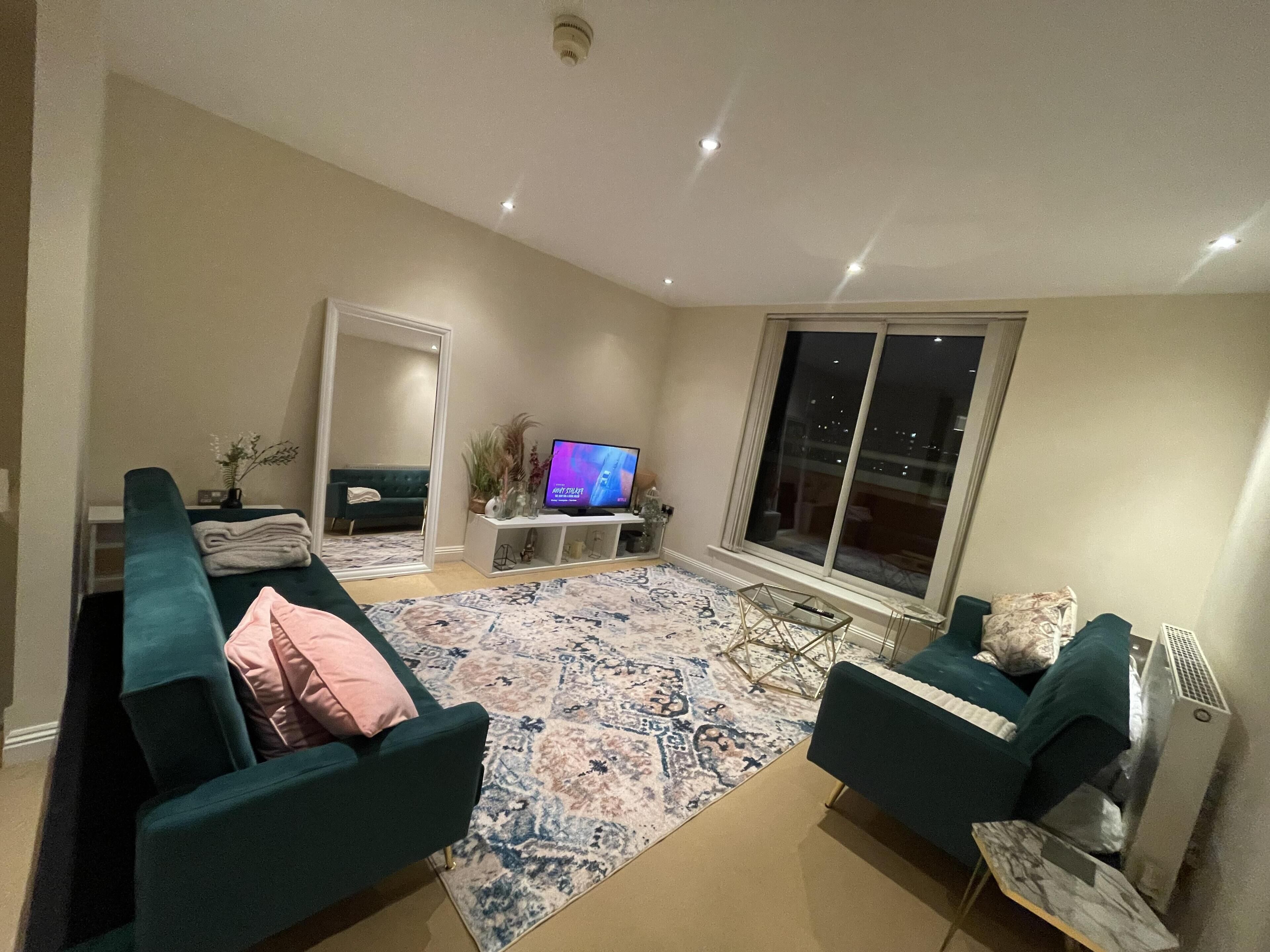 Un salon moderne avec des canapés de couleur turquoise, un tapis à motifs, un téléviseur à écran plat et une grande fenêtre.