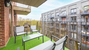 Un balcon avec une table en verre et deux chaises, un mur en briques et, au fond, un immeuble moderne doté de balcons.