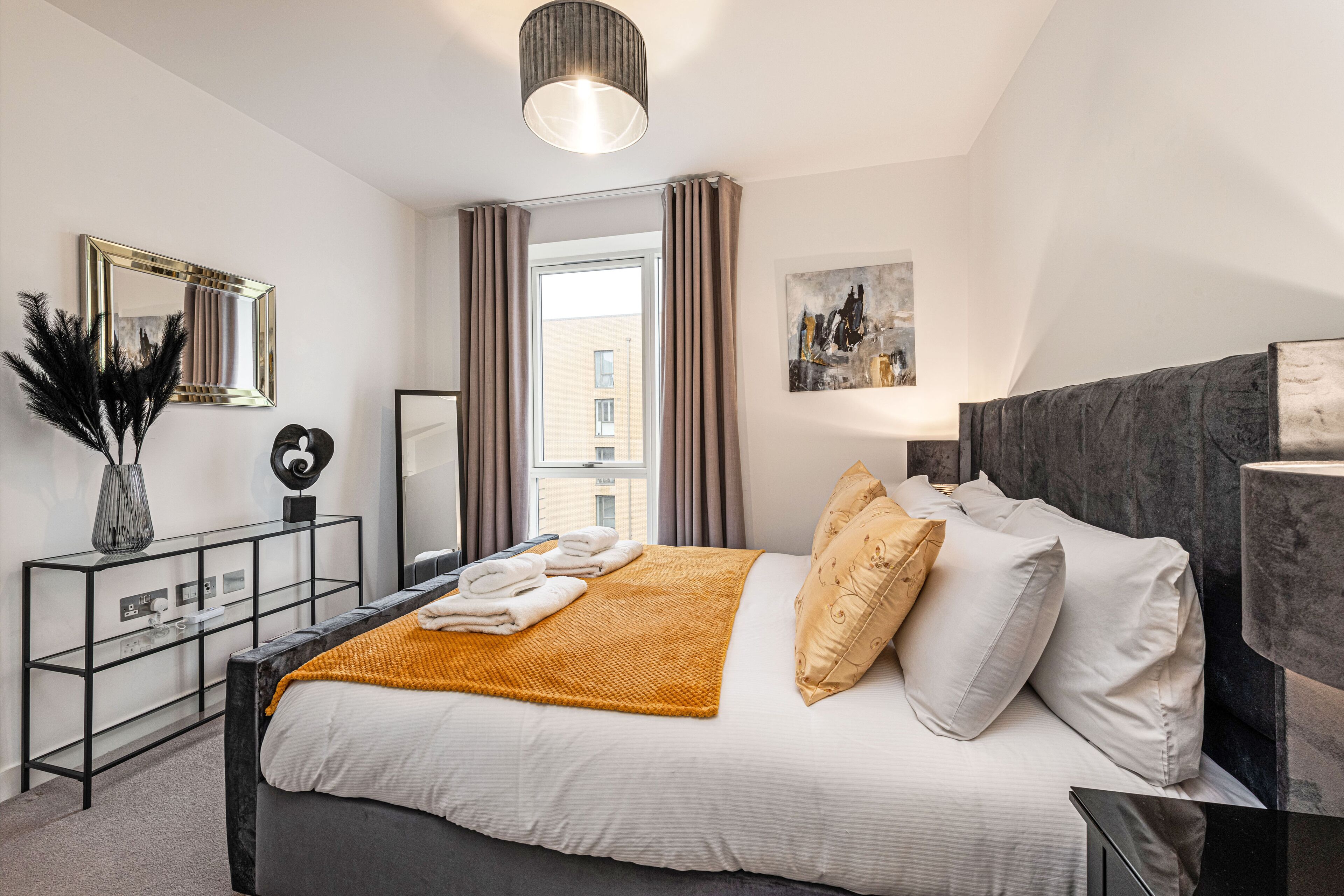 A modern bedroom with a large bed, a bedside table, a mirror, and a window with curtains.