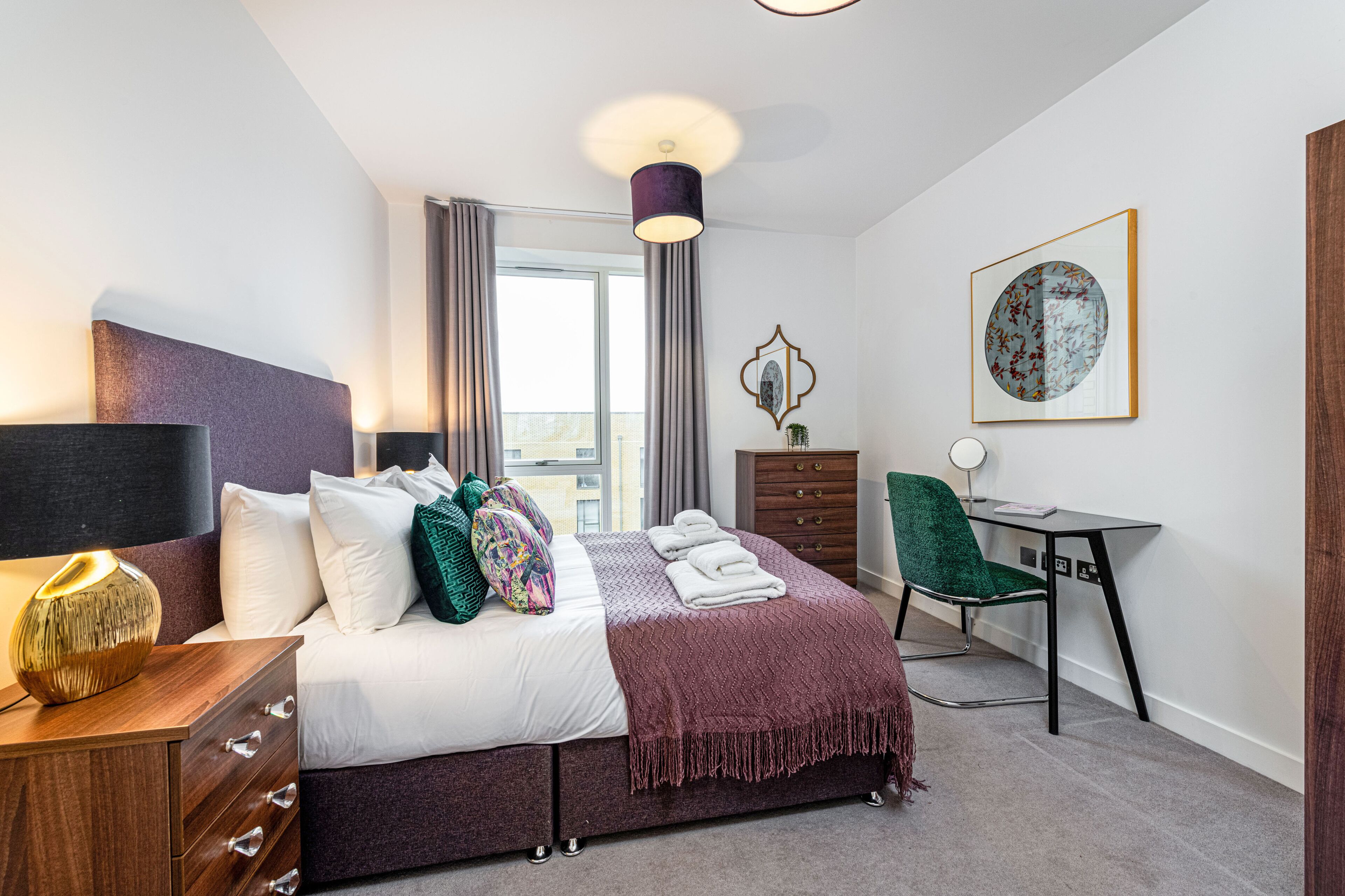 A modern bedroom with a bed, bedside table, dresser, desk, chair, and a large window with curtains.