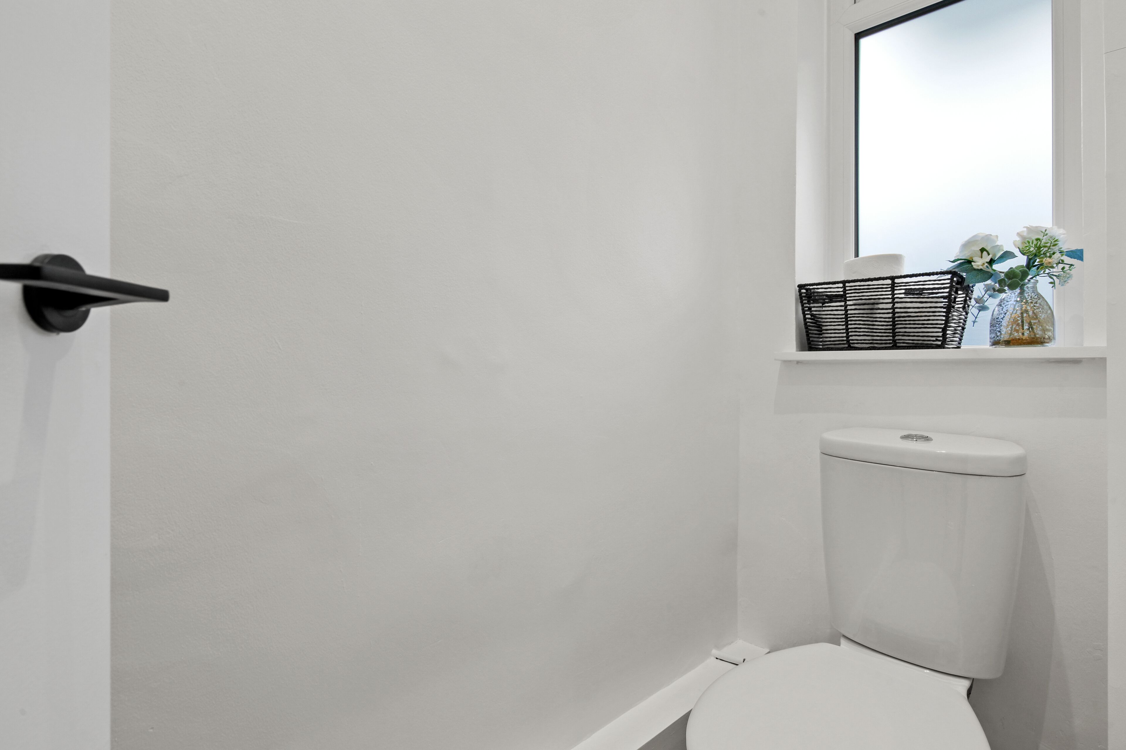 Ein helles Badezimmer mit Toilette, einem kleinen Fenster mit Spiegel und einem Regal mit Korb und Blumen in einer Vase.