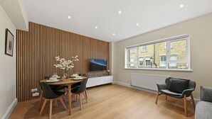 Un salon moderne avec une table à manger en bois, des chaises noires, un canapé gris et une grande fenêtre donnant sur des immeubles.