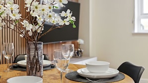 Une table à manger avec de l’assiette blanche, des bols et des verres à vin, un vase avec des fleurs blanches et un téléviseur en arrière-plan.