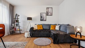 Una sala moderna con un sofá seccional negro, una mesa redonda, una lámpara de pie, una estantería y un amplio ventanal con cortinas traslúcidas.