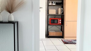 Un pasillo estrecho con suelo de baldosas blancas, una consola negra con un jarrón de plantas secas, un mueble negro con electrodomésticos de cocina y un armario color beige.
