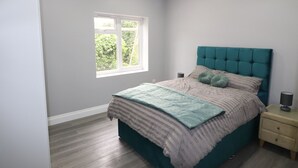 A bedroom with a teal headboard, a bed with striped bedding, a window with a view of greenery, and a bedside table with a lamp. - Impeccable 1-bed Apartment in Dartford (Dartford)