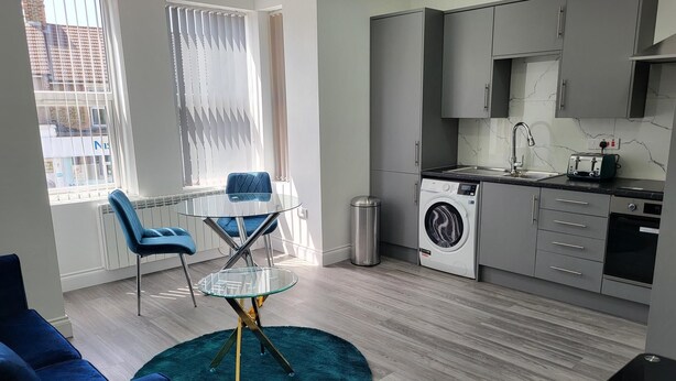 A modern kitchen with grey cabinets, a built-in washing machine, and a small dining area with a glass table and blue chairs. - Impeccable 1-bed Apartment in Dartford (Dartford)