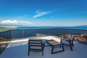 Un balcon doté d’une balustrade en verre, de deux chaises, d’une table et offrant une vue sur une ville côtière et la mer.
