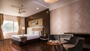 A hotel room with a large bed, a desk with two chairs, a ceiling fan, and a window with curtains. - HOTEL AADRIKA (Chikkamagaluru)