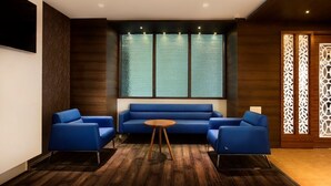 A modern room with a blue sofa set, a round wooden table, and a large window with frosted glass panels. - HOTEL AADRIKA (Chikkamagaluru)