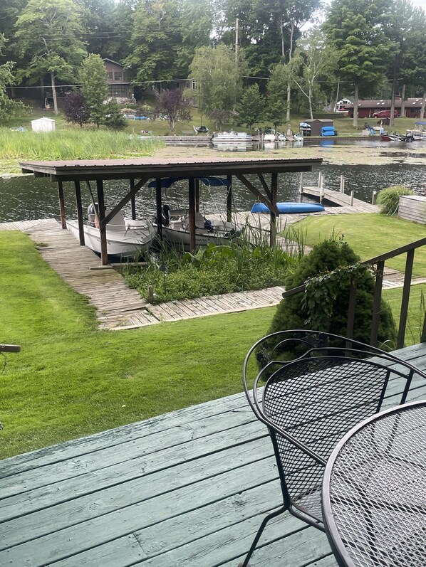 A wooden deck with a covered seating area, a small boat docked at the pier, and a grassy lawn with a view of a lake and houses in the background.