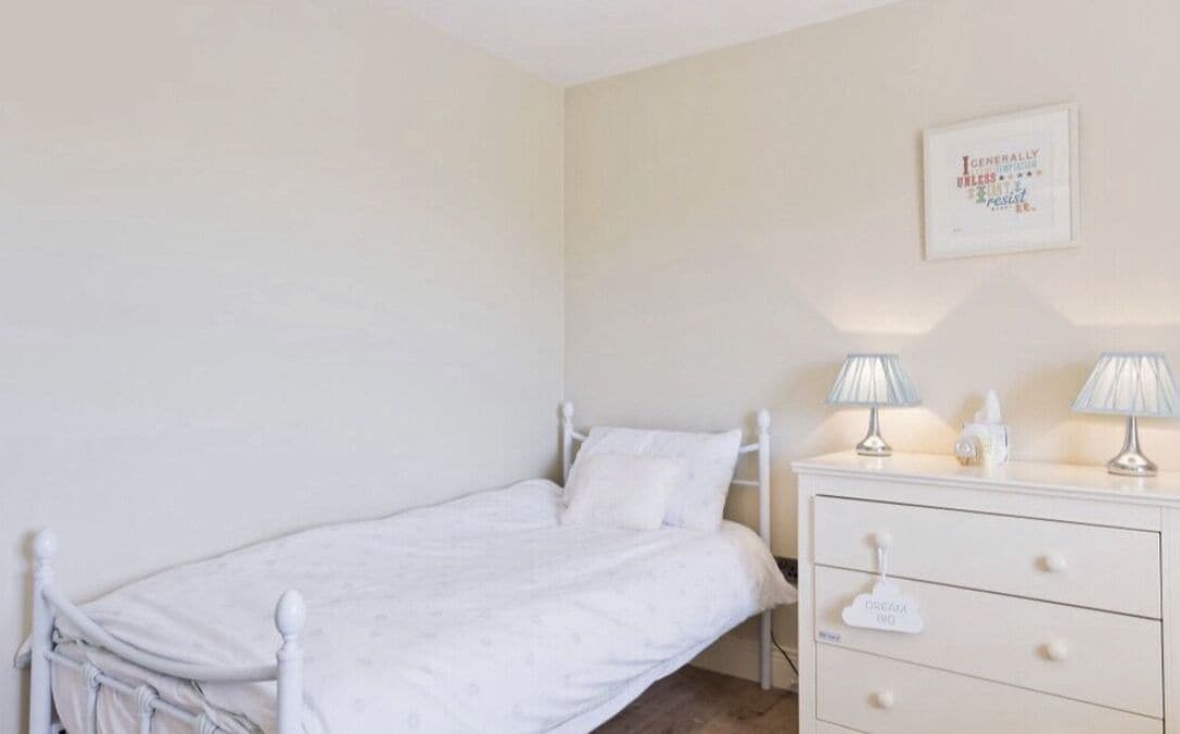A small, neatly made bedroom with a single bed, a bedside table with lamps, and a dresser with drawers.