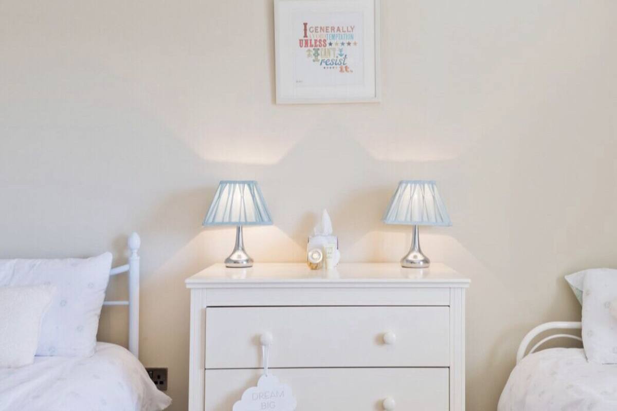 Un dormitorio ordenado con una cama blanca, una mesita de noche con dos lámparas y una cita inspiradora enmarcada en la pared.