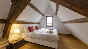 Een slaapkamer met een schuine plafond, een bed met witte beddengoed, rode kussens, een bedlampje en een klein raam.