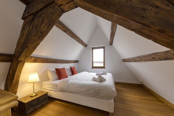 Un dormitorio con techo inclinado, cama con ropa de cama blanca, almohadas rojas, una lámpara de noche y un ventanal pequeño.