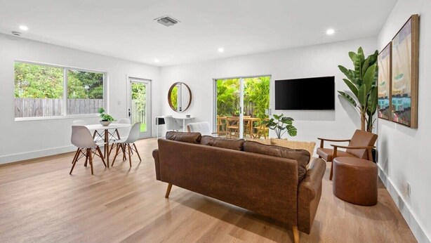 A spacious living room with a brown sofa, dining table, and chairs. There are large windows, a flat-screen TV, and a plant. - Fresh & Bright Condo 1BR/1BA – Near Riverwalk (Fort Lauderdale)