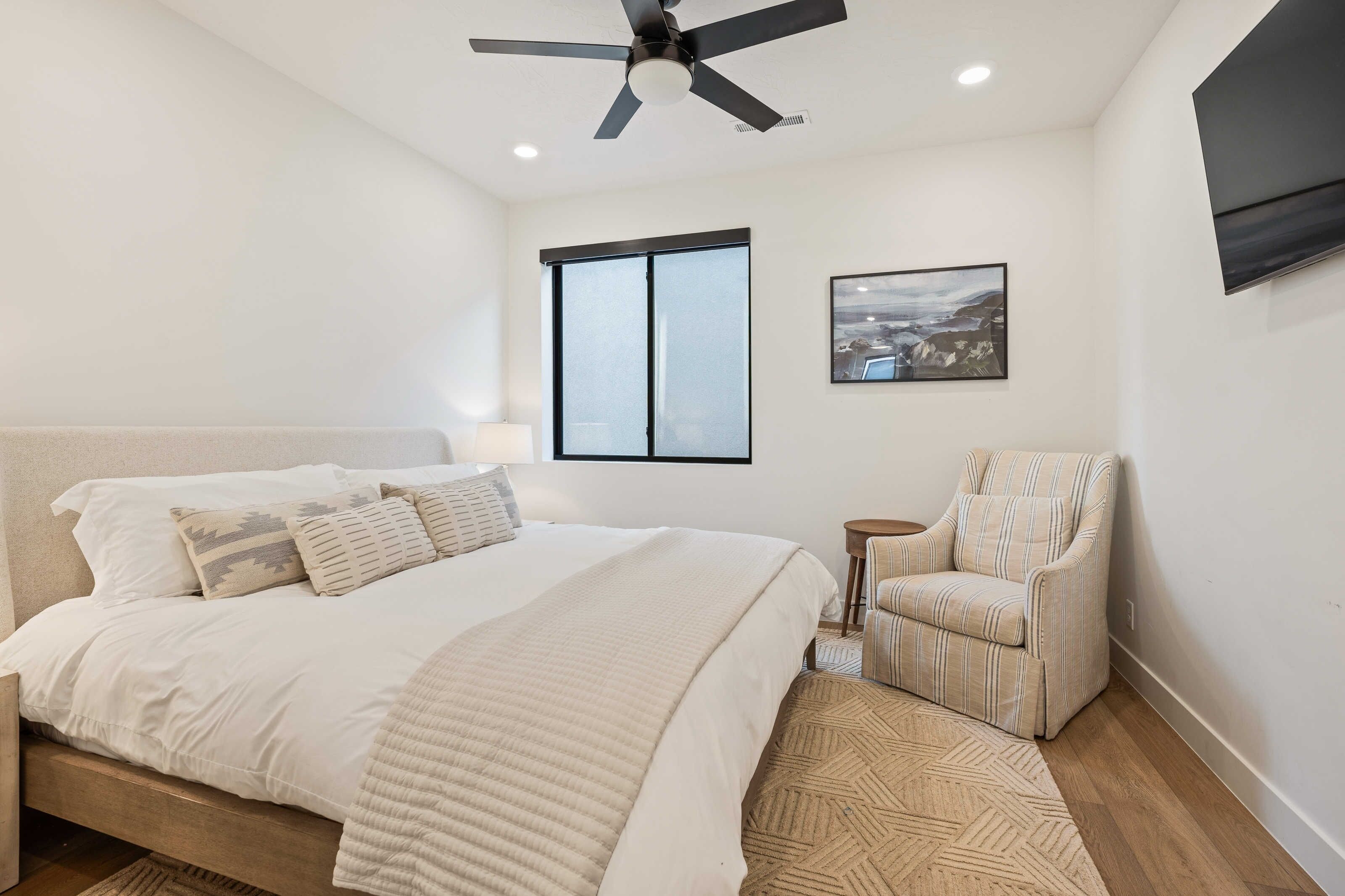 Quarto com cama, cadeira, ventoinha no teto, janela e um quadro na parede.
