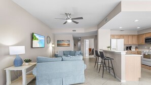 A modern living room with a sofa, a coffee table, and a TV. There is a kitchen area with a refrigerator and a microwave. - Crown Colony #101 - 16146 Mount Abbey Way 2 Bedroom Condo by RedAwning (Fort Myers)