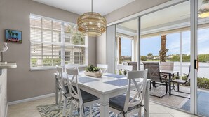 A dining area with a white table, blue placemats, and white chairs. There is a large window with blinds, a hanging light fixture, and a view of a body of water. - Crown Colony #101 - 16146 Mount Abbey Way 2 Bedroom Condo by RedAwning (Fort Myers)
