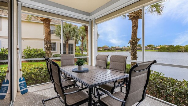 A covered patio with a table and chairs, large windows, and a view of a lake and palm trees. - Crown Colony #101 - 16146 Mount Abbey Way 2 Bedroom Condo by RedAwning (Fort Myers)