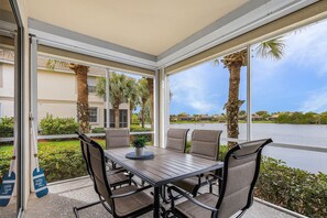 Un patio techado con mesa y sillas, ventanas amplias y vista a un lago y palmeras.