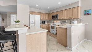 A modern kitchen with wooden cabinets, a central island, a microwave, and a stove. - Crown Colony #101 - 16146 Mount Abbey Way 2 Bedroom Condo by RedAwning (Fort Myers)
