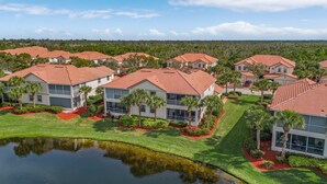 A residential area with multiple houses featuring red-tiled roofs and white walls, surrounded by lush greenery and a small body of water. - Crown Colony #101 - 16146 Mount Abbey Way 2 Bedroom Condo by RedAwning (Fort Myers)