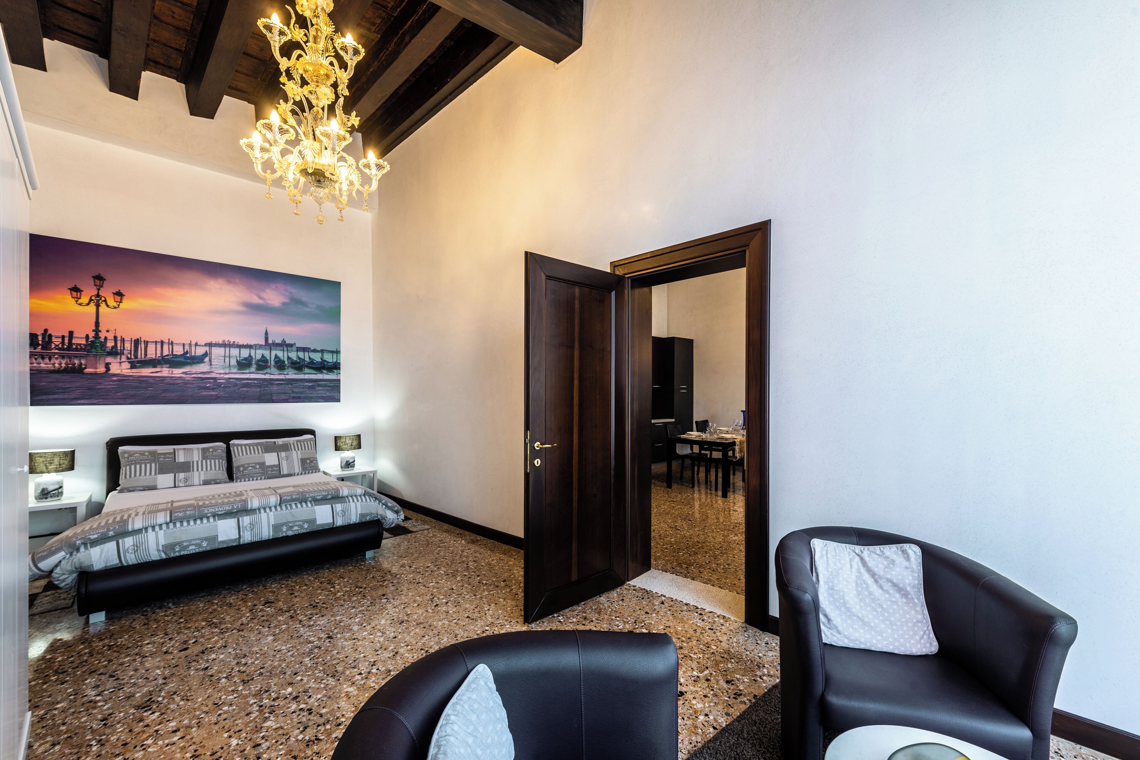 A bedroom with a bed, a painting, a chandelier, and a door leading to another room.