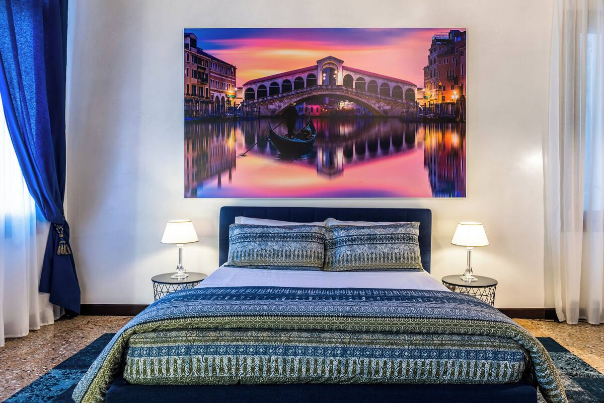 A hotel room with a large bed, two bedside tables, a nightstand, a painting of a bridge, and a view of the room's interior.
