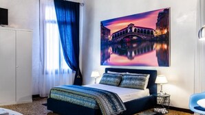 A hotel room with a bed, a nightstand, a lamp, a chair, a window with curtains, and a painting of a bridge. - Ca' Sant'Angelo (Venice)