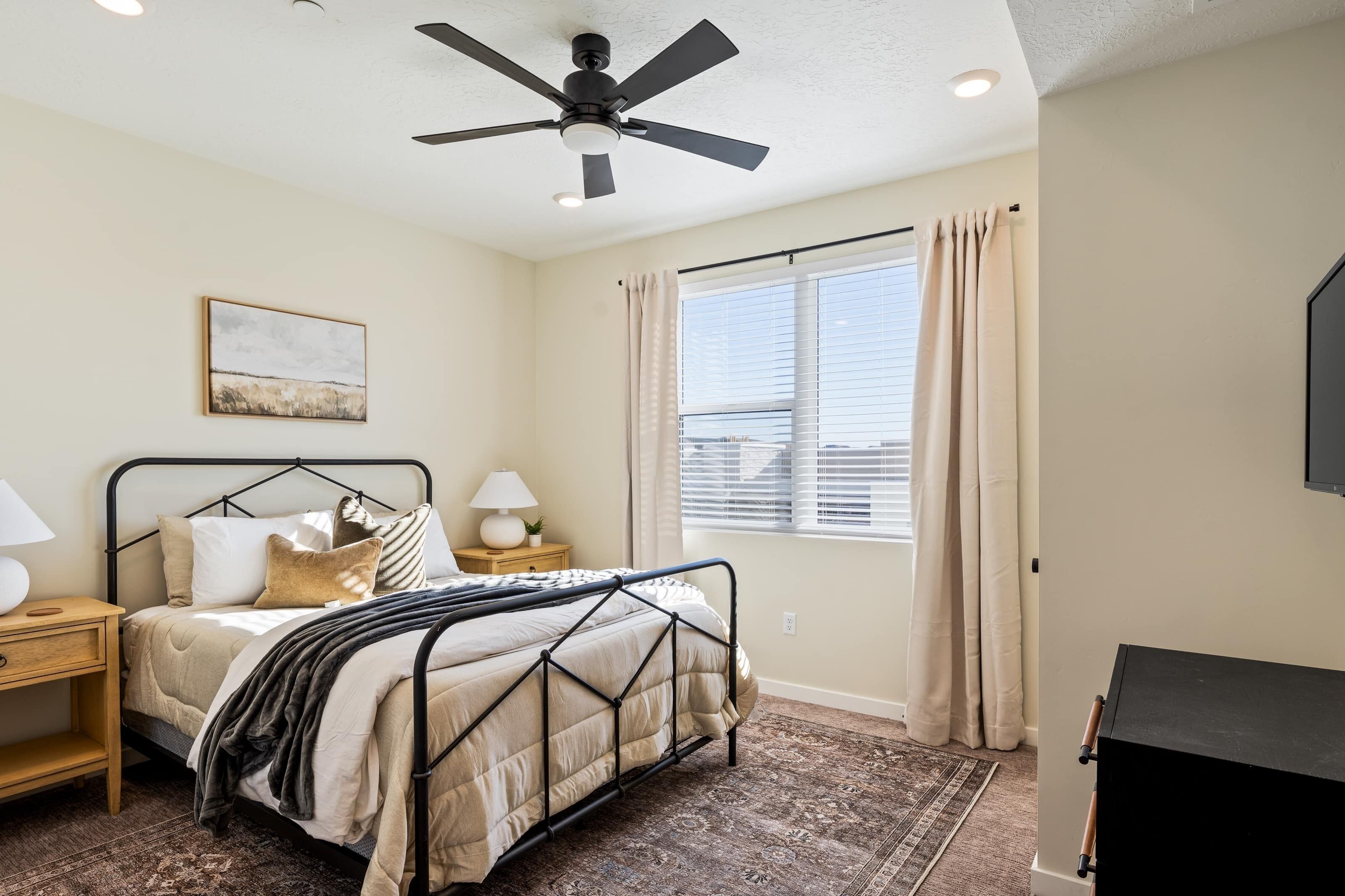 A bedroom with a bed, a ceiling fan, a window with curtains, a nightstand, a lamp, and a painting on the wall.