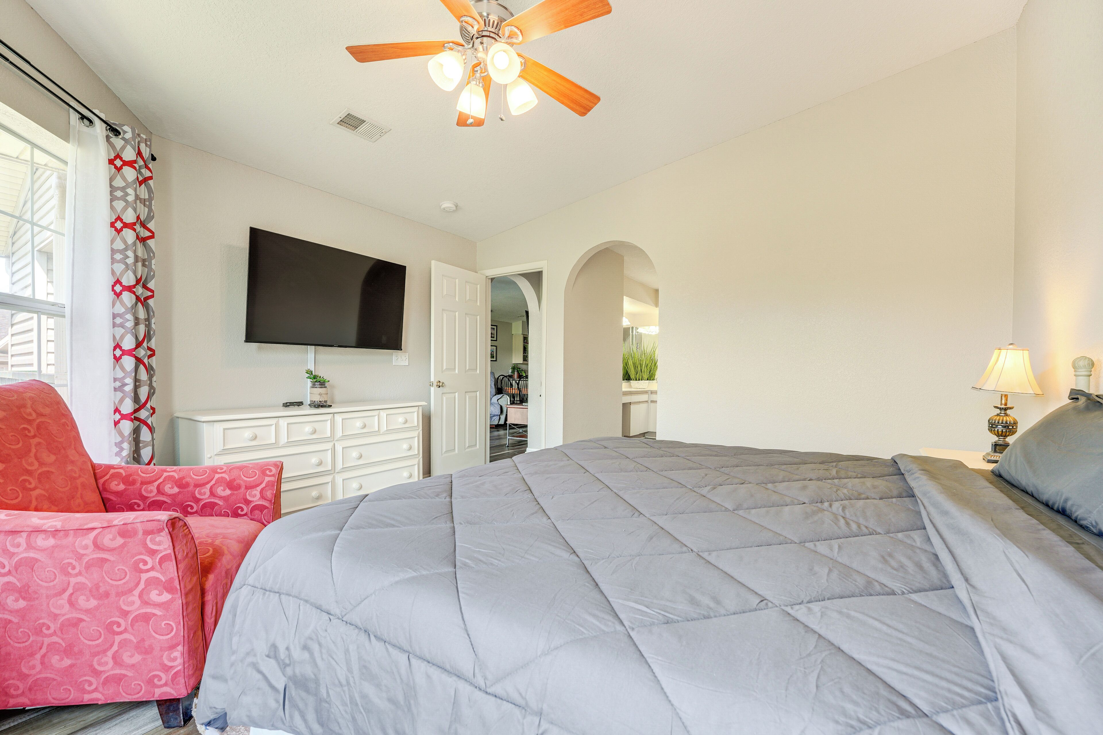 A bedroom with a large bed, a ceiling fan, a TV mounted on the wall, and a window with curtains.
