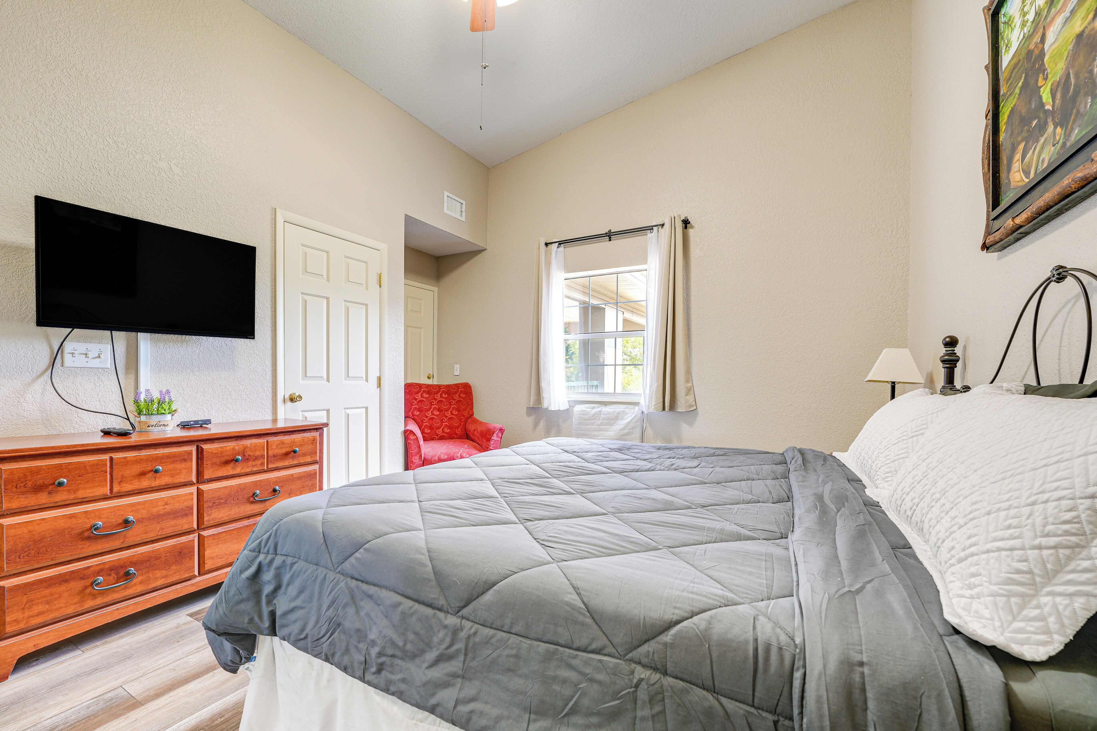 A bedroom with a bed, a dresser, a television, a window with curtains, and a painting on the wall.
