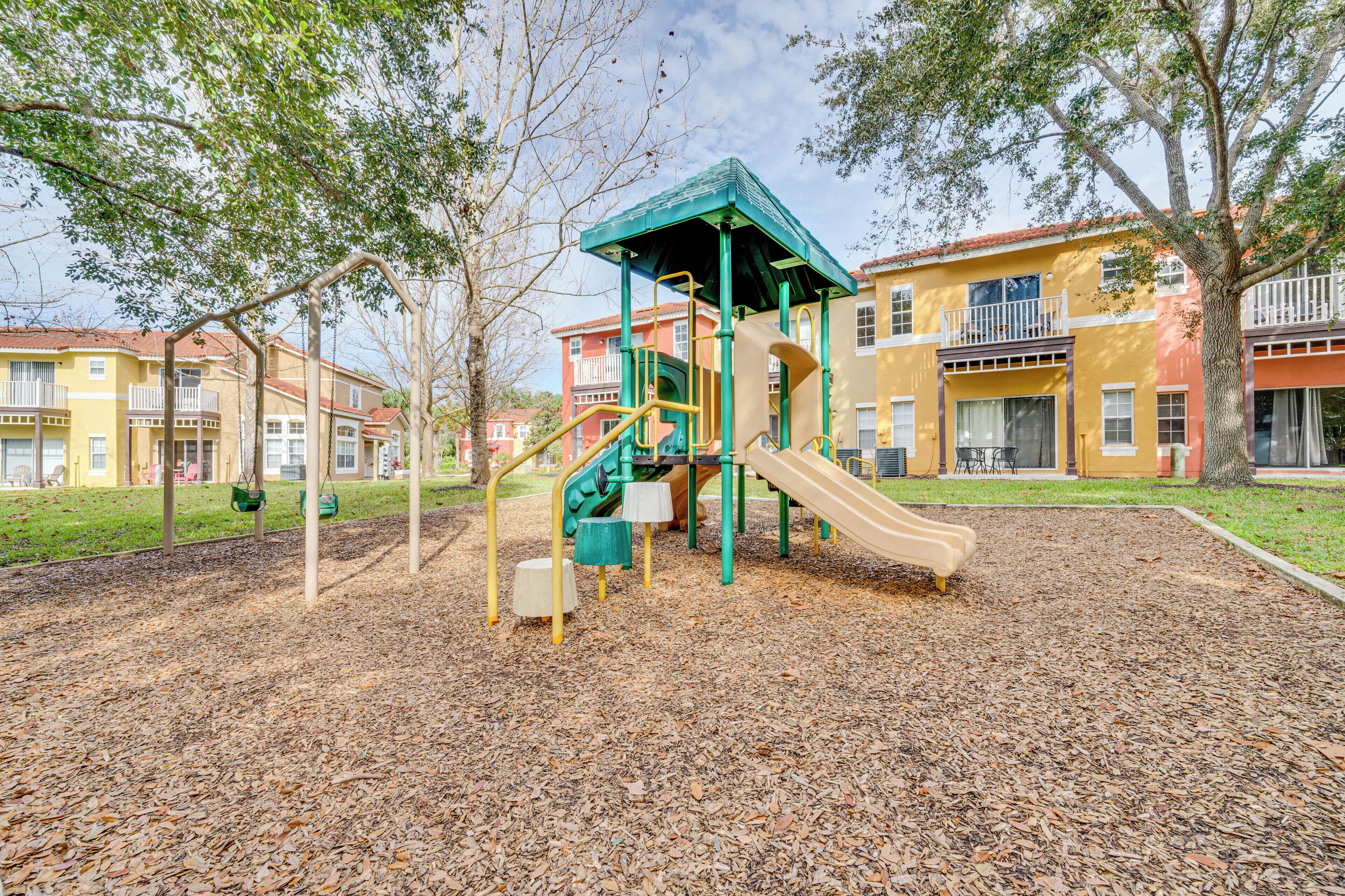 Un parque infantil con un tobogán y estructuras para escalar, rodeado de árboles y edificios residenciales.